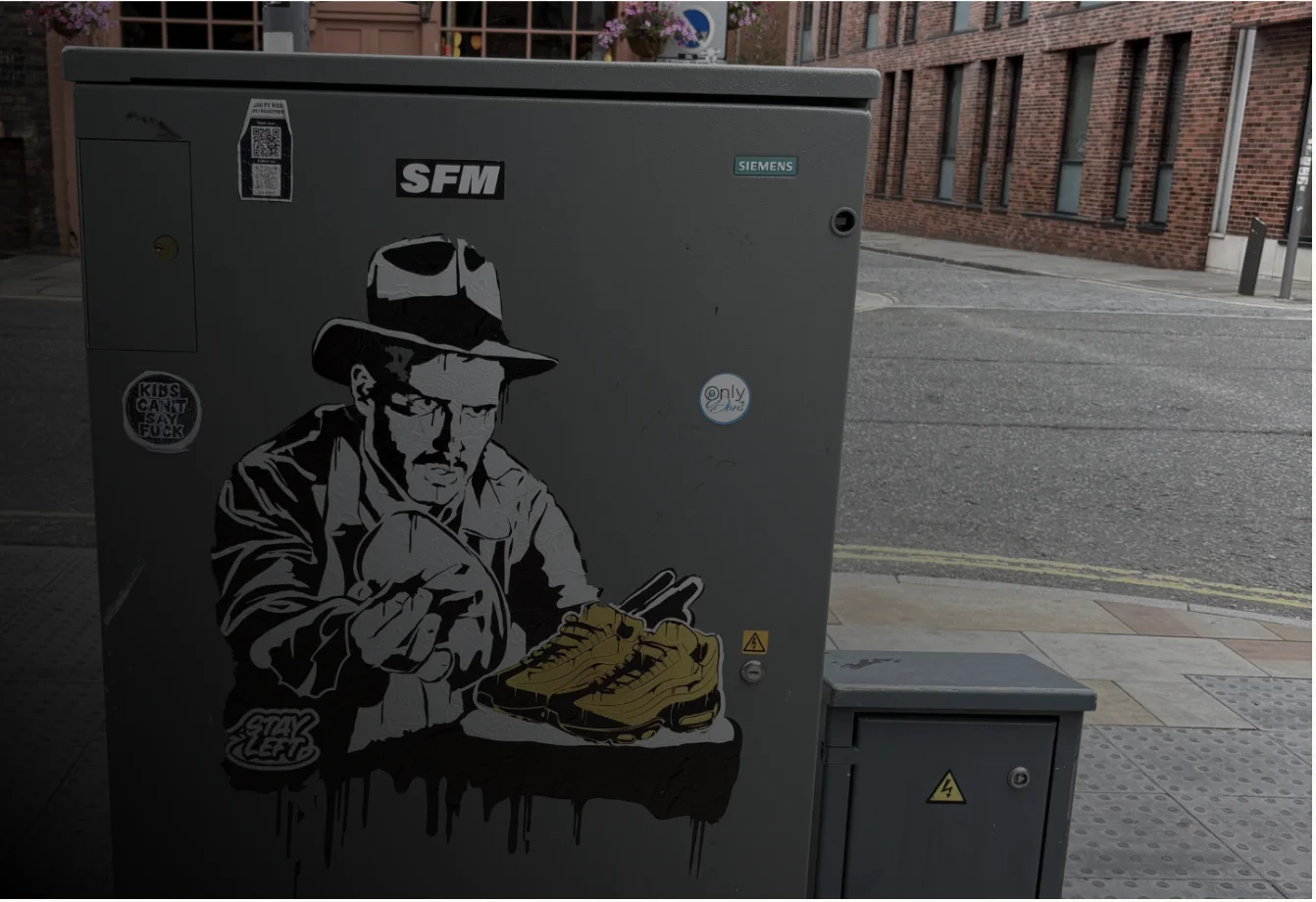 A street cabinet pasted with a stencil of a man in a hat and coat holding a shoe, beside a pair of yellow trainers