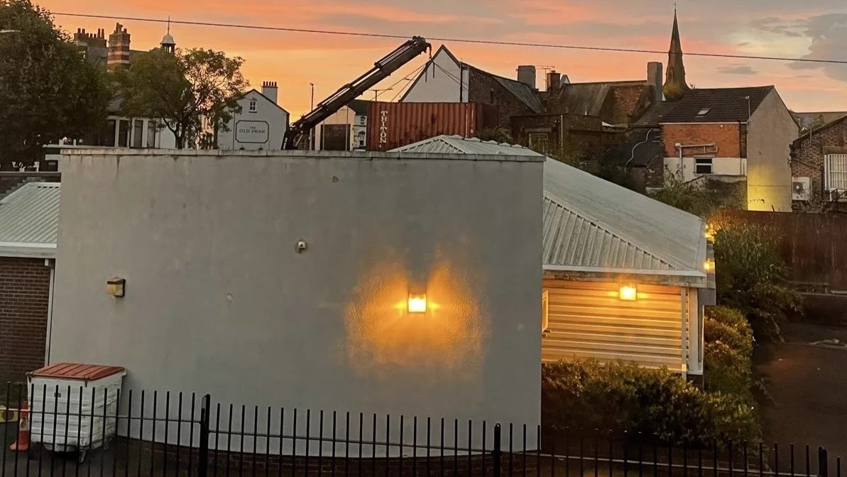 A white rendered building at dusk, its wall lit by a single lamp, with a crane, a shipping container and a church spire against an orange sky behind