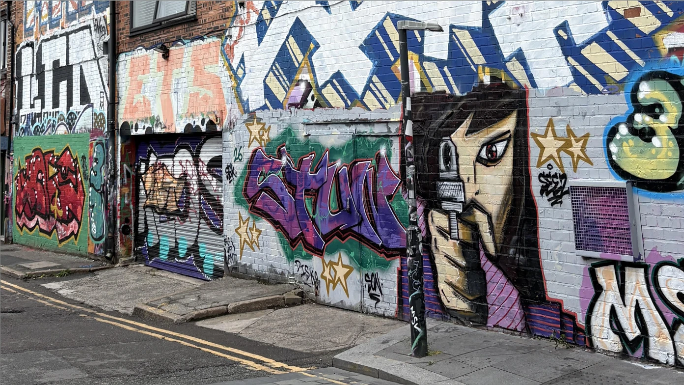 A back-street wall painted end to end with graffiti, including a large face holding a camera to one eye